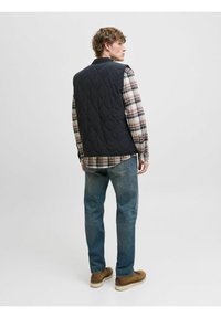 Marineblå quiltet vest med høj krave, båret over en ternet skjorte. Standard pasform med en glat tekstur, parret med blå jeans og brune sko.