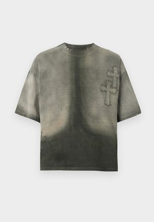 Πλυμένο, σκούρο πράσινο oversized T-shirt με φθαρμένα υφασμάτινα σταυρωτά μπαλώματα στο στήθος και κοντά στο κάτω αριστερό μέρος.