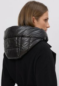 Czarny wełniany płaszcz z błyszczącym czarnym, puchowym kapturem. Kaptur ma pikowaną teksturę z widocznymi detalami przeszyć.