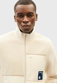 Beige Fleecejacke mit hohem Kragen, Reißverschluss und Fronttasche. Mit einer strukturierten Oberfläche und einem blauen Logo-Patch auf der Tasche.