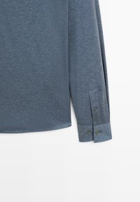Camicia a maniche lunghe di colore blu scuro, realizzata in tessuto testurizzato. Presenta polsini con bottone con accenti in azzurro a contrasto e orlo arrotondato.