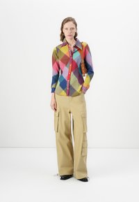Camicia a maniche lunghe colorata con motivo a rombi in tonalità di rosso, giallo e blu; abbinata a pantaloni cargo beige con tasche laterali.