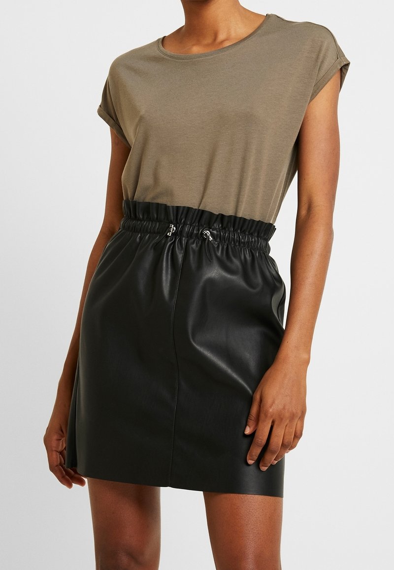 Femme portant un haut taupe à manches courtes rentré dans une jupe en cuir noire taille haute avec une ceinture élastique froncée.