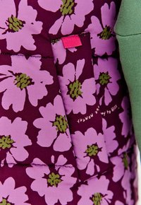 Materiał pikowany w duże fioletowe kwiaty z zielonymi środkami, z naszytą różową metką i częściowo widocznym zielonym rękawem na krawędzi.