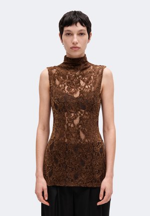 Le mannequin se tient face à l’objectif, vêtu d’un haut en dentelle marron sans manches, d’un pantalon noir à jambes larges et de bottines noires, sur un fond clair uni.