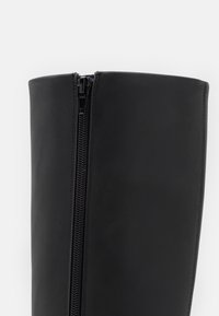 Bota de piel negra hasta la rodilla con una cremallera visible en el lateral, textura suave y diseño estructurado. Sin adornos adicionales.