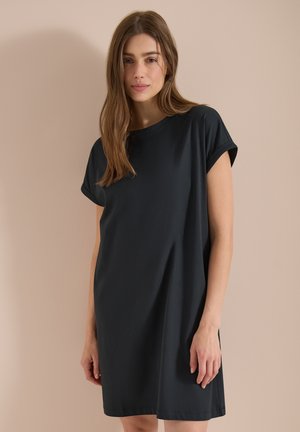 Jeune femme aux longs cheveux bruns portant une robe noire ample à manches courtes sur un fond beige.