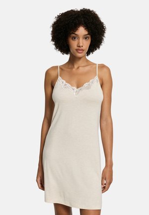 Vrouw die een beige mouwloze slipjurk draagt met kanten rand bij de halslijn, staand tegen een effen lichte achtergrond.