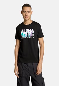 Musta puuvillapaita, jossa on värikäs "ALPHA INDUSTRIES" -logo aaltoilevilla yksityiskohdilla. Lyhyet hihat ja pyöreä pääntie.