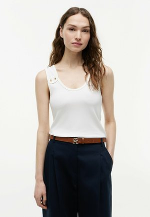 Kobieta o długich falowanych włosach, ubrana w biały bezrękawnik z guzikami na ramiączku oraz ciemne spodnie z wysokim stanem i brązowym paskiem.