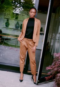 Brauner maßgeschneiderter Blazer und passende Hose mit elastischem Bund, getragen über einem schwarzen Rollkragenpullover. Schwarze Stiletto-Absätze vervollständigen den Look.