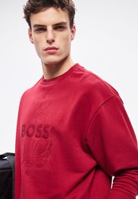 Το κόκκινο φούτερ διαθέτει ανάγλυφο λογότυπο "BOSS" και υφή με λεπτομέρειες. Έχει στρογγυλή λαιμόκοψη και ριμπ μανσέτες, κατασκευασμένο από λείο ύφασμα.