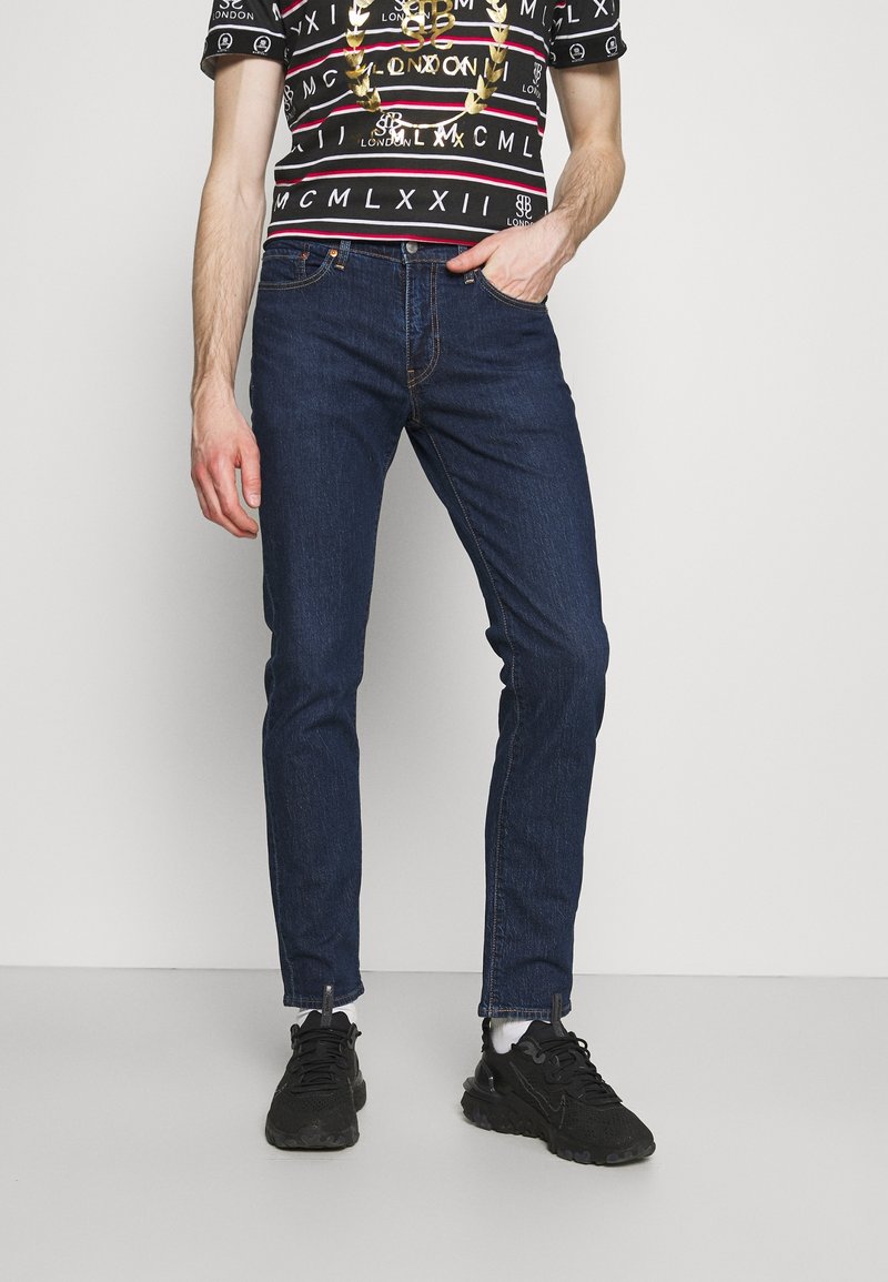 Jean en denim bleu foncé à coupe slim. Porté avec un t-shirt noir à motifs et des baskets noires. Texture lisse avec des coutures standard.