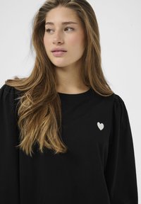 Schwarzes, langärmliges Oberteil aus weichem Stoff mit rundem Ausschnitt und einem kleinen weißen Herzmotiv auf der linken Brust.