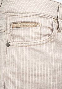 Jeans rayés beige avec une finition texturée, présentant une étiquette de marque marron foncé sur la poche arrière et des accents en métal.