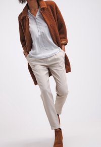 Texturerad brun kappa över en ljus mönstrad blus, kombinerad med beige byxor och bruna ankelstövlar för en lager-på-lager-look.