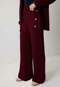 Cilvēks ar bordo adījuma platām biksēm un atbilstošu pogu kardigānu, apakšā valkājot beigtas slaidas kurpes, stāv uz baltas grīdas.