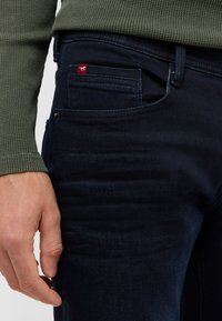 Ciemnoniebieskie dżinsy o slim fit z czerwonym logo na tylnej kieszeni i subtelną fakturą. Ręka oparta na udzie.