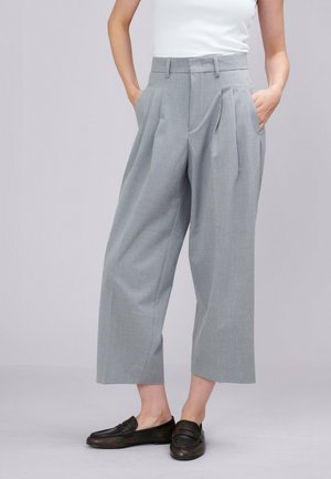 Vrouw draagt lichtgrijze wijde cropped broek met voorplooien en zwarte loafers, handen in de zakken, wit topje zichtbaar.