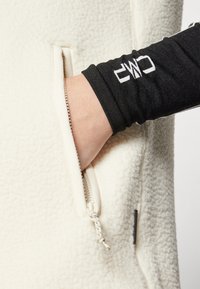 Cremfarbener Fleece-Zip-Up-Jacke mit Seitentasche. Verfügt über eine schwarze, langärmlige Unterlage. Der Reißverschluss hat eine Stoffschlaufe für einfachen Gebrauch.