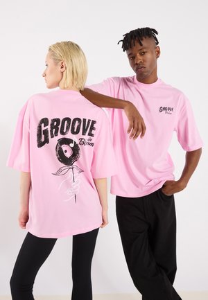 Lyserøde oversized t-shirts med sort grafisk tryk; forsiden har "Groove" logo, bagsiden har et design med en hånd, der holder en rose.