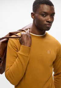 Felpa gialla senape con scollatura tonda e polsini a coste. Una borsa in pelle marrone è appesa alla spalla, accentuando il look informale.