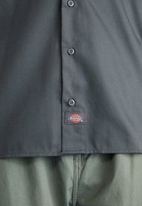 Chemise gris foncé à boutons avec une étiquette avec le logo Dickies près du bas, portée sur un pantalon vert clair.