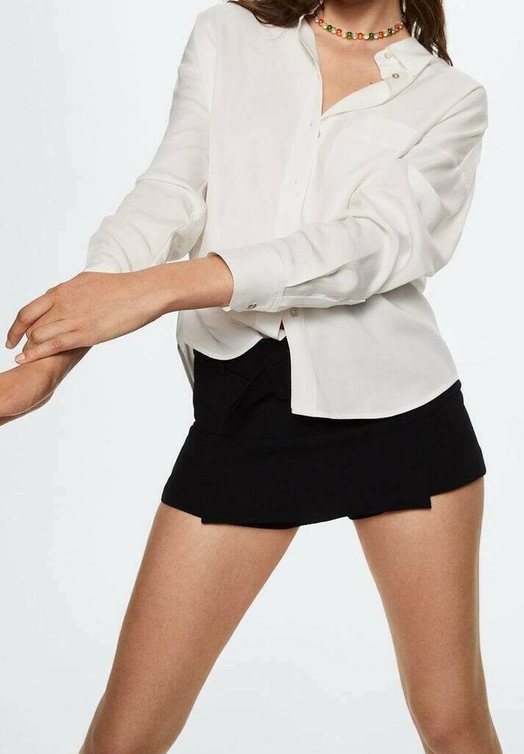Femme portant une chemise blanche boutonnée et une mini-jupe noire, ajustant les manches avec les bras croisés devant son corps.