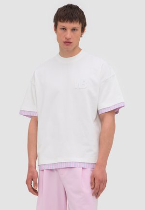 Young man wearing a white oversized T-shirt over a pink and blue striped shirt, paired with light pink pants, standing against a plain background.