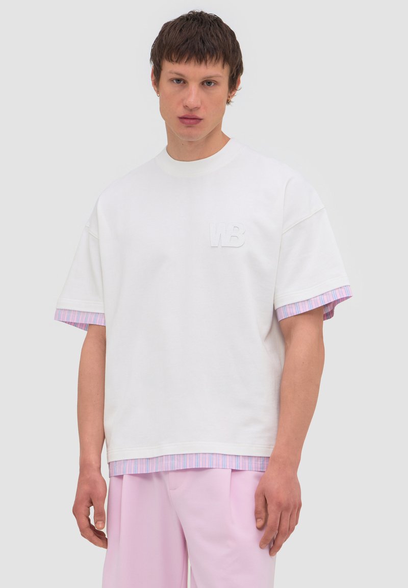 Νέος άνδρας που φοράει ένα λευκό oversized T-shirt πάνω από ένα ροζ και μπλε ριγέ πουκάμισο, συνδυασμένο με ανοιχτό ροζ παντελόνι, στέκεται μπροστά από ένα μονόχρωμο φόντο.
