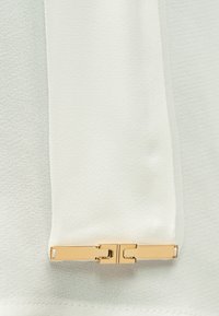 Λευκό ύφασμα με ομαλή υφή και λεπτομέρεια αντίθετου χρυσού μεταλλικού κούμπωματος, που αναδεικνύει έναν καθαρό, μινιμαλιστικό σχεδιασμό.