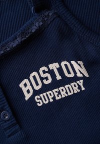 Marineblauwe geribbelde tanktop met witte tekst "BOSTON SUPERDRY". Voorzien van een kantafwerking langs de halslijn en een klein knoopdetail.