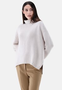 Hellbeiger Strickpullover mit hohem Kragen, lässiger Passform und seitlichen Schlitzen; kombiniert mit beigen Hosen, die einen strukturierten Stoff zeigen.