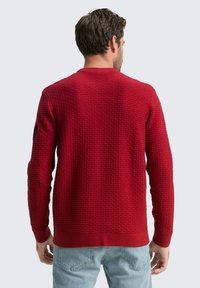 Rode textuurtrui met een ronde hals, ribbels bij de mouwen en de zoom. Platte breipatroon zichtbaar, gedragen over lichtblauwe jeans.