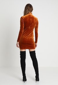Samtiges, rostfarbenes Minikleid mit langen Ärmeln und hohem Kragen, kombiniert mit kniehohen schwarzen Wildlederstiefeln und Stöckelschuhen.