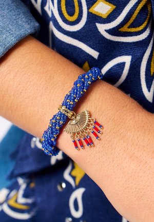 Bracelet tressé bleu avec des accents dorés et un pendentif en forme de éventail, muni de perles rouges et bleues suspendues ; fond textile visible.