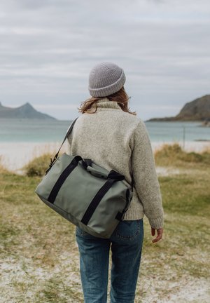 Person med grått strikket lue og genser, bærer en grå sportsbag over skulderen, går mot en strand med fjell i bakgrunnen.