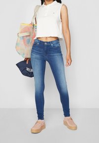 Camisa polo blanca ajustada, jeans azules ceñidos y zapatos de plataforma rosa claro. La modelo sostiene una bolsa de tote colorida y una gorra azul marino.