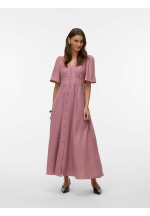 Donna che indossa un abito lungo color malva con bottoni e maniche corte a sbuffo, indossando tacchi neri appuntiti, in piedi davanti a uno sfondo bianco uniforme.