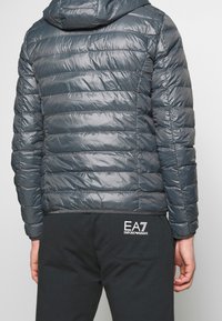 Polstrovaná šedá bunda s lesklým povrchem, zdobená vodorovným prošíváním, s připevněnou kapucí a elastickým lemem. Nošena s černými kalhotami, viditelné logo.