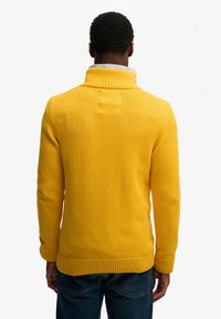 Żółty dziergany sweter z wysokim kołnierzem, ściągaczem na dole i mankietach, z centralnym szwem na plecach i fakturowanym wykończeniem.