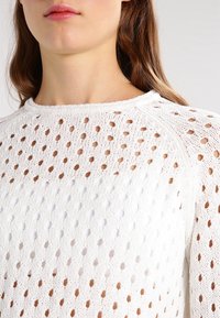 Pull à mailles blanches présentant des motifs ajourés, un col arrondi et des accents côtelés au col et aux poignets. Tissu léger et texturé.