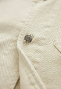 Primo piano di un tessuto di velluto a coste beige con un prominente bottone in metallo argentato con testo e un pattern testurizzato ben visibile.