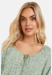 Ljusgrön blus med blommönster, veck, rund halsringning med nyckelhålsknyte och strukturerad tyg för extra detalj.