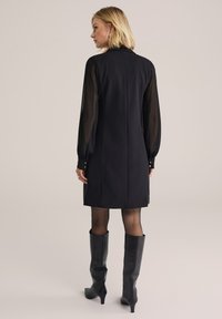 Rochie neagră cu guler înalt și mâneci vaporoase, bufante. Prezintă un corset ajustat, croi drept și accente cu nasturi la manșete. Botine negre până la genunchi.