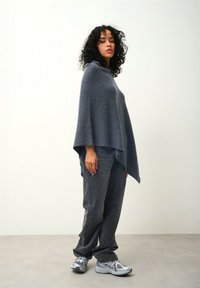 Poncho grigio carbone realizzato in tessuto morbido, caratterizzato da una vestibilità comoda e aperture laterali, abbinato a jeans grigi e scarpe sportive argentate.