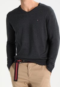 Pull gris foncé à manches longues avec col en V, en matière douce, arborant un petit logo sur la poitrine. Associé à un pantalon beige et une ceinture rayée.