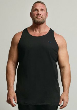 Muskulöser Mann mit Bart steht frontal, trägt ein schwarzes ärmelloses Tanktop mit kleinem Logo auf der Brust, neutraler Hintergrund.