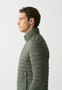 Vihreä puhvijakku, jossa on vaakasaumastus, korkea kaulus ja vetoketjusulku. Vasemmassa hihassa on logopatch.