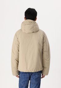 Beige Daunenjacke mit hohem Kragen und strukturiertem Stoff, mit cinched Saum und weiten Ärmeln, kombiniert mit blauen Jeans.